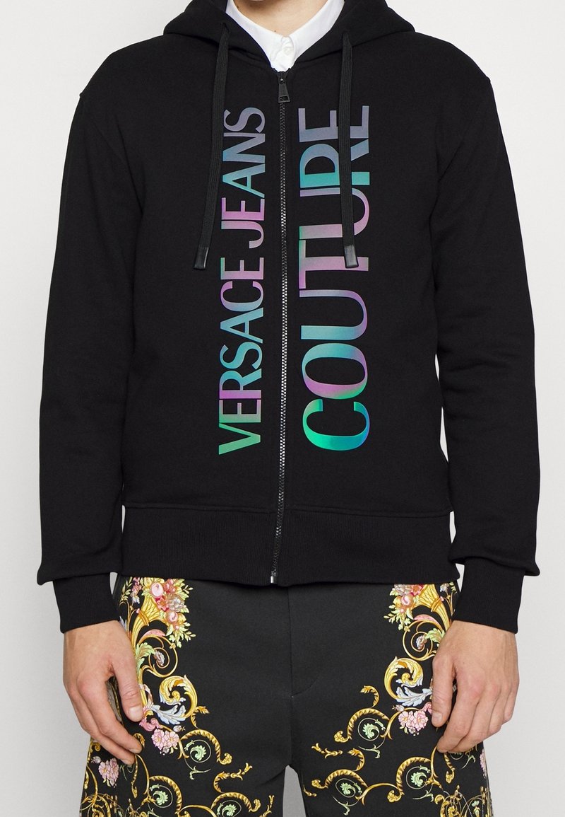 Sweat à capuche noir zippé avec un logo multicolore "Versace Jeans Couture" sur le devant, porté avec un pantalon noir présentant des motifs baroques dorés et floraux ornés.