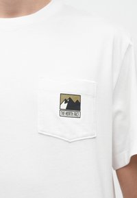 Λευκό βαμβακερό T-shirt με τσέπη που φέρει μαύρο και χρυσό λογότυπο βουνού. Στρογγυλή λαιμόκοψη και κοντά μανίκια. Ομαλή υφή.