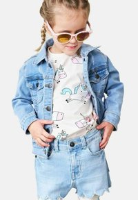 Vaaleansininen farkkutakki ja aaltohelmaiset shortsit, yhdistettynä valkoiseen paitaan, jossa on pastellinvärisiä yksisarvisia. Vaaleanpunaiset aurinkolasit viimeistelevät asun.