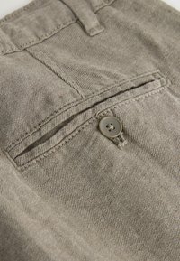 Gros plan sur un tissu beige montrant une poche arrière boutonnée avec des coutures détaillées et une boucle de ceinture au-dessus.