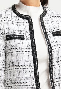 Giacca di tweed bianca con cuciture nere, rifiniture nere e tasche. Il tessuto presenta un motivo testurizzato a griglia.