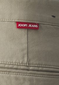 Beige cotton fabric with stitched details and a red label reading "JOOP! JEANS" in white text.