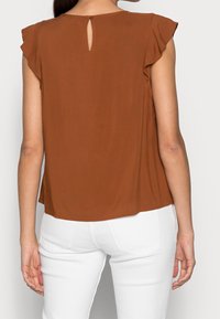 Camisa marrón de manga corta con abertura en la parte posterior, hecha de una tela suave, combinada con pantalones blancos para crear contraste.