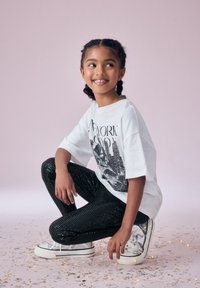 T-shirt grafico bianco con skyline di una città, abbinato a leggings neri con paillettes e sneakers argentate. Lo sfondo è rosa chiaro con coriandoli.