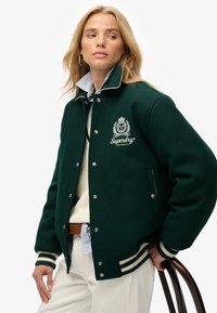 Groene wollen jas met contrasterende witte gestreepte manchetten en kraag. Heeft een logo op de borst en zijzakken. Model draagt casual kleding.
