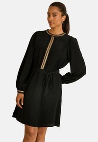 Schwarzes Kleid mit langen Ärmeln und gerafften Schultern, das mit einem goldenen Besatz entlang der Vorderseite und einem gebundenen Taillengürtel für eine definierte Silhouette versehen ist.