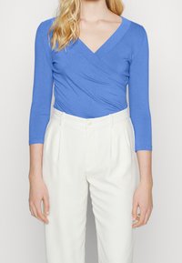 Mujer vistiendo una blusa ajustada azul estilo envolvente con mangas de tres cuartos y pantalones blancos de talle alto con pliegues.