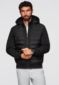 Giacca nera imbottita con cappuccio, apertura con zip e polsini a costine. Presenta una texture liscia e pannelli in tessuto a contrasto.