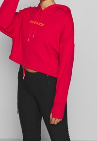 Sudadera recortada roja con cordones y texto amarillo "FIRE + ICE". Combinada con pantalones negros que tienen bolsillos con cremallera. Textura de tela suave.