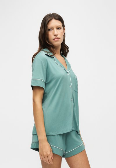 Femme portant un haut turquoise à manches courtes boutonné et un short assorti avec un liseré blanc, debout sur un fond blanc uni.
