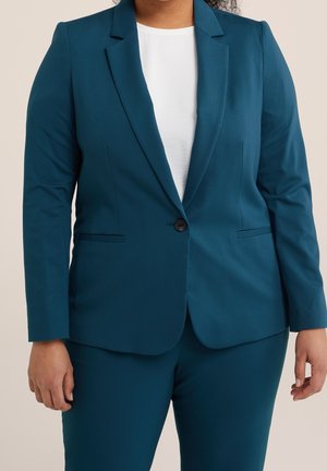 Kvinde iført turkis tætsiddende blazer med enkeltknapslukning og matchende bukser over en hvid top, stående mod en lys baggrund.