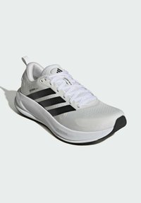 Zapatilla deportiva blanca con diseño de tres franjas negras, superficie texturizada, parte superior de malla y suela de goma, que cuenta con un cuello acolchado y cordones.