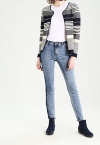 Gebreid vest met navy en crèmekleurige strepen, gecombineerd met een wit t-shirt en lichtblauwe skinny jeans. Enkelhoge zwarte laarzen maken de outfit compleet.