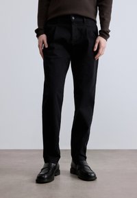 Juodos medvilninės kelnės su tiesiu kirpimu ir priekinėmis klostėmis, su sagų užsegimu; dėvimos su juodais mokasinai, ant neutralaus fono.