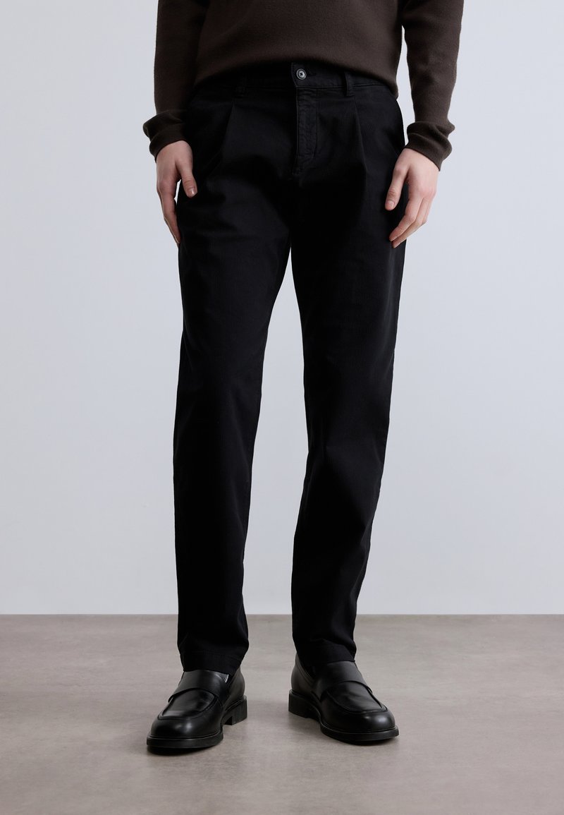 Juodos medvilninės kelnės su tiesiu kirpimu ir priekinėmis klostėmis, su sagų užsegimu; dėvimos su juodais mokasinai, ant neutralaus fono.