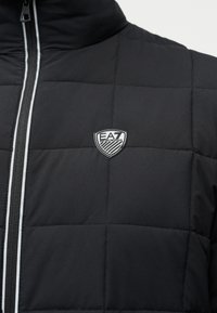 Μαύρο καπιτονέ μπουφάν με ψηλό γιακά, κλείσιμο με φερμουάρ και ασημένιο λογότυπο με ρίγες κατά μήκος του φερμουάρ. Λείο υλικό.