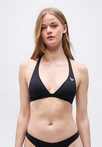 Νεαρή γυναίκα που φοράει μαύρο μαγιό μπικίνι με λουράκι στον λαιμό και μικρό λογότυπο στην αριστερή πλευρά, στέκεται μπροστά σε έναν απλό φωτεινό φόντο.