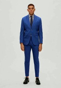 Modrá saková bunda a nohavice so slim fit strihom, s pruhovanou košeľou a čiernou kravatu, doplnené čiernymi formálnymi topánkami.