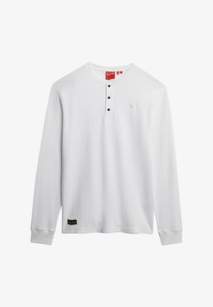 T-shirt henley blanc à manches longues avec trois boutons noirs, col rond et petite étiquette de marque noire près de l'ourlet.
