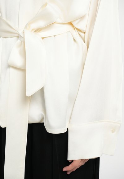 Blusa in seta crema con cintura annodata, tessuto plissettato, maniche oversize e polsini abbottonati, abbinata a un sottogiacca nera.