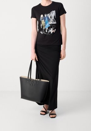 Femme portant un t-shirt graphique noir avec deux femmes et des chiens, jupe noire froncée, sandales noires à brides, tenant un grand sac fourre-tout DKNY noir.