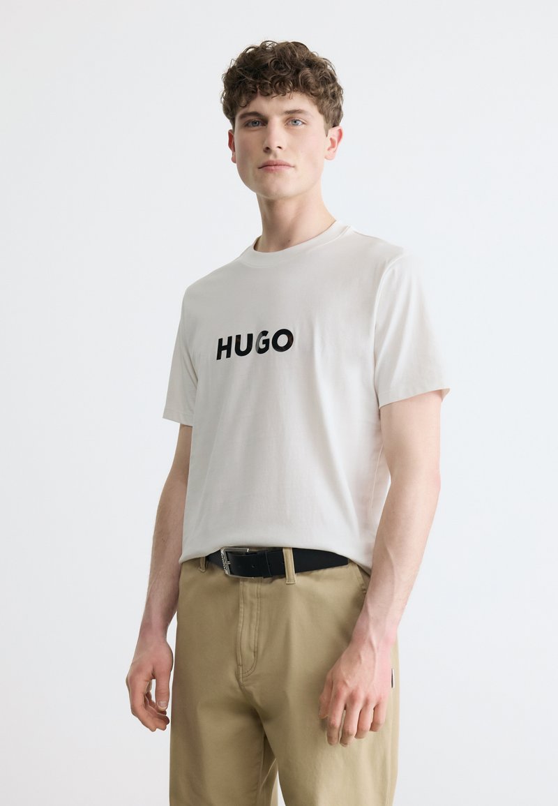 Νεαρός άνδρας με κοντά σγουρά μαλλιά, που φοράει λευκό T-shirt με το κείμενο "HUGO" και μπεζ παντελόνι, μπροστά σε απλό λευκό φόντο.