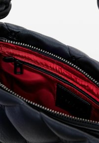 Interno della borsa in pelle nera con una morbida fodera in tessuto rosso, zip argento e un piccolo etichetta nera. Dettagli di cucitura texturizzati visibili.