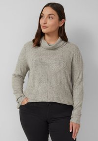Frau mit schulterlangem braunem Haar, die einen grauen strukturierten Rollkragenpullover und schwarze Hosen trägt, die Hände in den Taschen, seitlich blickend.