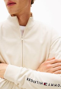 Veste crème zippée avec un col haut et des poignets côtelés. Présente le logo "Tommy Hilfiger" en bleu marine le long de la manche. Texture douce et lisse.
