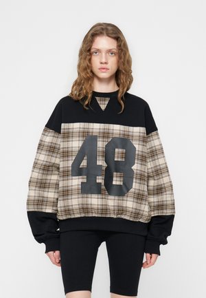 Jeune femme aux cheveux ondulés portant un sweat-shirt oversize à carreaux et noir avec le numéro 48 et un short de cyclisme noir, debout devant un fond blanc.