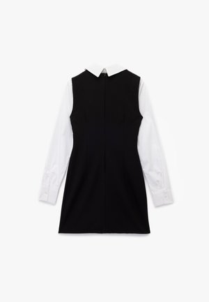 Musta hihaton mekko valkoisen pitkähihaisen paidan päällä, jossa on terävä kaulus ja napilliset kalvosimet, nähtynä takaa.