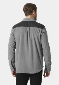 Szara polarowa koszula z czarnymi akcentami na ramionach, długimi rękawami i zaokrąglonym dołem. Posiada zapięcia na guziki przy mankietach oraz miękką teksturę.