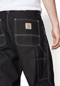 Pantalones cargo negros en denim duradero, con costuras blancas y una etiqueta cuadrada de Carhartt con un logo amarillo en el bolsillo trasero.