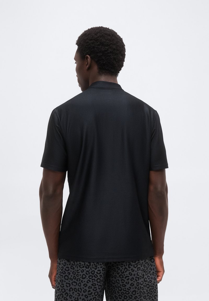 Homme portant une chemise noire à manches courtes et un short foncé à imprimé léopard, debout, dos tourné vers le spectateur sur un fond blanc.