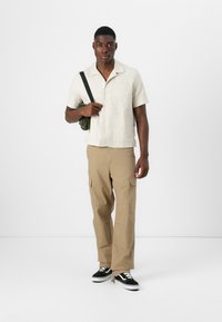Camisa corta beige claro de manga corta, con botones y bolsillo en el pecho; pantalones cargo marrones con bolsillos laterales; zapatillas negras y blancas; llevando una bolsa verde.