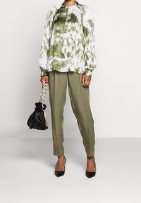 Osoba w oliwkowej bluzce z długim rękawem w kolorze tie-dye, oliwkowych spodniach tapered, czarnych szpilkach, trzymająca czarną torbę z dużym łańcuchem jako uchwytem.