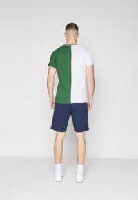 Man standing with back to camera wearing a green and white split short-sleeve shirt, navy shorts, white socks, and white sneakers.