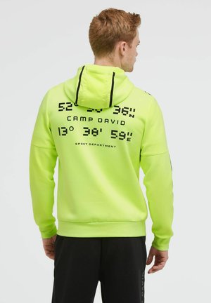 Man wearing bright yellow hoodie with black text and coordinates on back, paired with black pants, facing away from the camera.