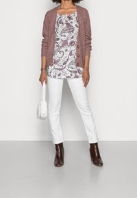 Kvinna som bär en brun stickad cardigan över en blommig topp, vita jeans, bruna ankelstövlar och håller en vit handväska, stående på ett grått golv.