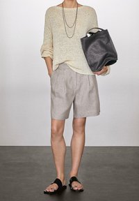 Osoba ubrana w beżowy sweter z dzianiny, jasnoszare szorty, czarne sandały, naszyjniki w warstwach i trzymająca dużą czarną skórzaną torbę.