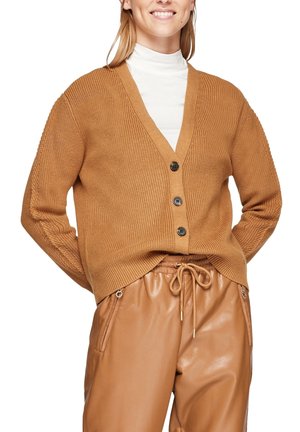 Femme portant un cardigan en maille marron boutonné sur un col roulé blanc, associé à un pantalon en cuir beige à cordon de serrage avec poches zippées.