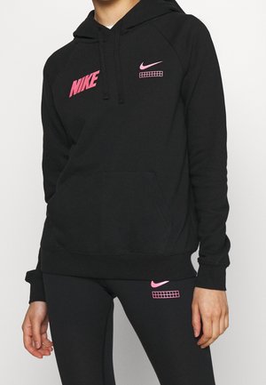 Osoba ubrana w czarną bluzę z kapturem i legginsy Nike z różowymi logo i graficznymi detalami na piersi i udzie, stoi na jednolitym tle.