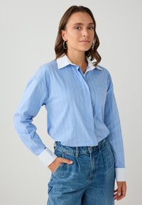 Vrouw met bruin haar die zilveren ringoorbellen draagt, een blauw gestreept shirt met witte kraag en manchetten, en hooggemonteerde blauwe jeans, staande met haar hand in haar zak.