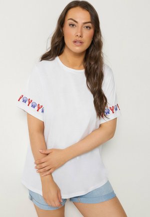 Frau mit langen braunen Haaren, die ein weißes T-Shirt mit roten Hummern und blauen Muscheln auf den Ärmeln sowie hellblaue Jeansshorts trägt.