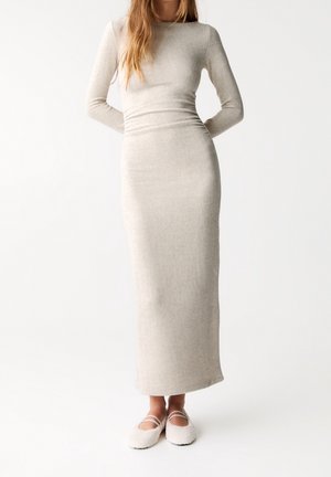 Kvinde iført en tætsiddende, langærmet, ankel-lang beige kjole med teksturerede cremefarvede tøfler, stående mod en ensartet hvid baggrund.