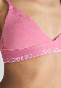 Roza trianglasta bralette z teksturiranim logotipom, ki ima kontrastni elastični pas z napisom "Calvin Klein" v beli barvi.