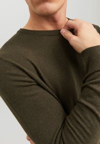 Olíva zöld kötött pulóver, kerek nyakkiv�ágással, bordázott mandzsettával és sima textúrával. Közeli felvétel az alkarjáról és kezéről, amint állítja a gallért.