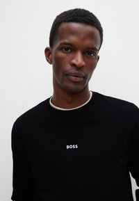 Svart T-skjorte med kort erm, rund hals og hvit "BOSS"-logo på brystet. Glatt tekstur, minimalistisk design.
