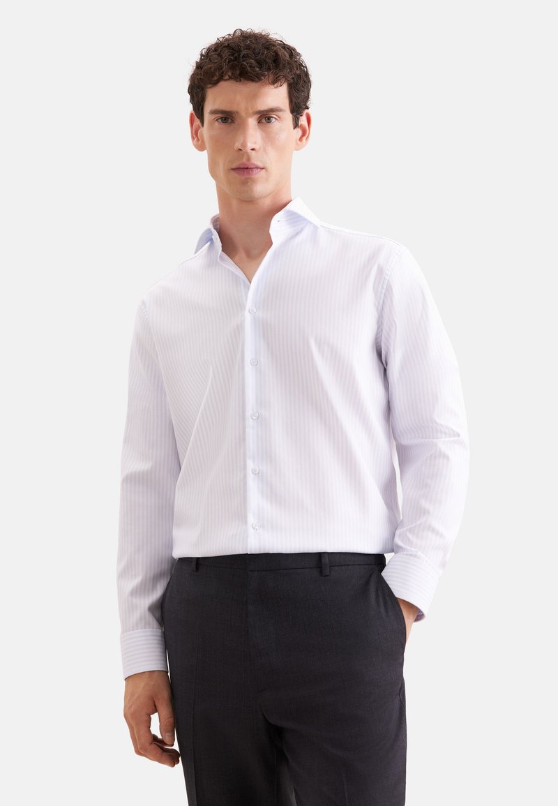 Homme portant une chemise blanche à manches longues avec des rayures discrètes et un pantalon de costume noir, debout avec une main dans la poche.