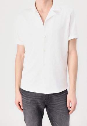 Personne portant une chemise blanche à manches courtes boutonnée et un jean gris foncé, debout devant un fond clair uni.